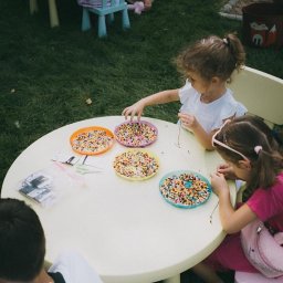 Kreatywne Urodziny Adriana Foryś - Dwie dziewczynki przy okrągłym stole na trawie, nawlekają kolorowe koraliki na nitki. Na stole cztery talerzyki z koralikami i woreczek ze sznurkami.