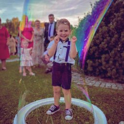 Kreatywne Urodziny Adriana Foryś - Uśmiechnięty chłopiec w szelkach stoi wewnątrz gigantycznej bańki mydlanej na trawie, w tle goście uroczystości. Kolorowe refleksy na bańce.