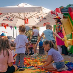 Kreatywne Urodziny Adriana Foryś - Dzieci bawią się kolorowymi patyczkami na kocu, w tle dmuchany zamek i animator pod parasolem z logo Żywiec. Impreza plenerowa dla dzieci.