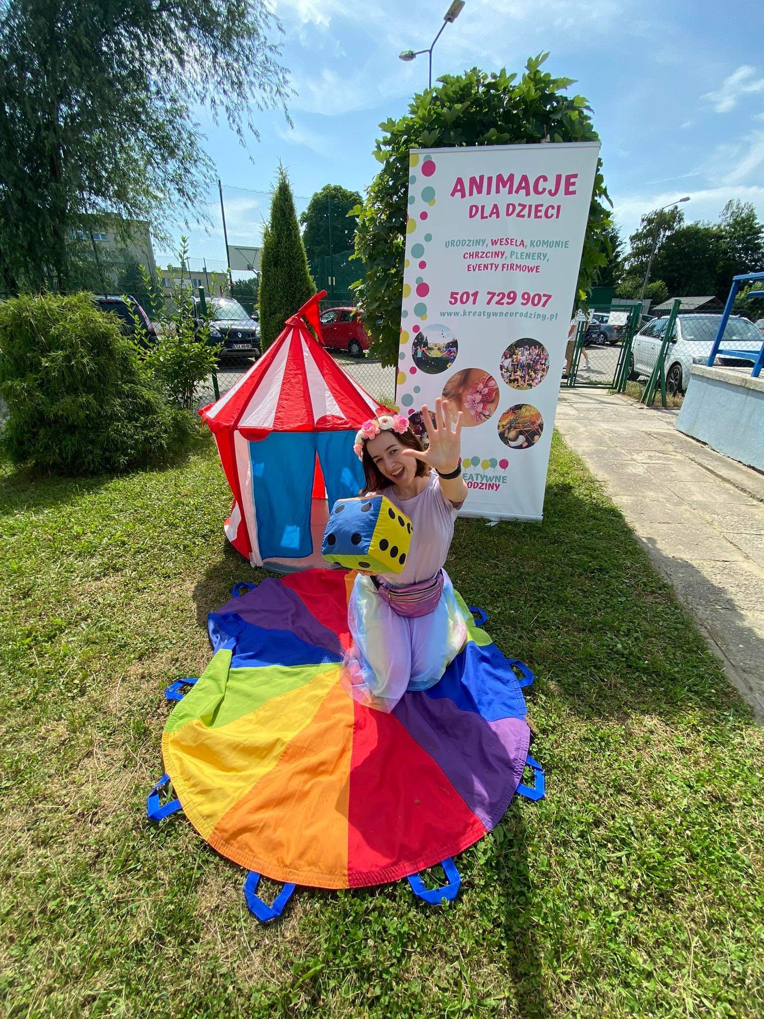 Uśmiechnięta animatorka w kostiumie, z kostką w dłoni, siedzi na kolorowej chuście. W tle namiot cyrkowy i baner reklamowy z napisem 'Animacje dla dzieci'.