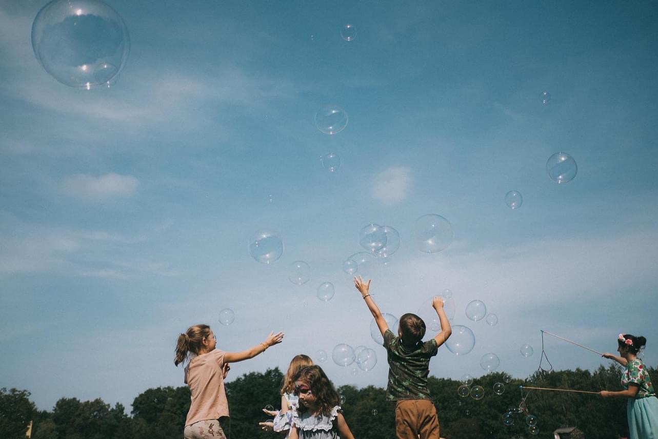 Dzieci bawią się bańkami mydlanymi na tle błękitnego nieba. Dziewczynka z boku puszcza bańki za pomocą sznurka i patyków. Radosna atmosfera zabawy na świeżym powietrzu.