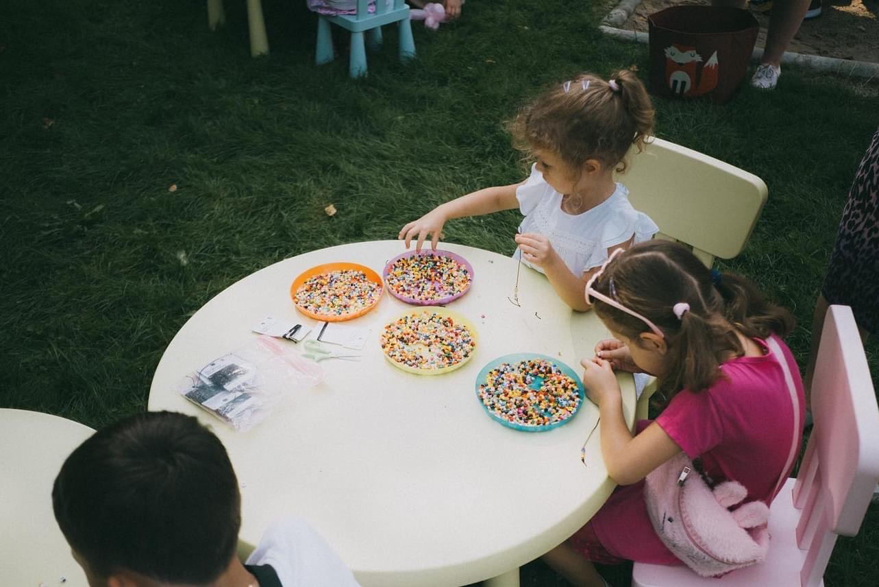 Dwie dziewczynki przy okrągłym stole na trawie, nawlekają kolorowe koraliki na nitki. Na stole cztery talerzyki z koralikami i woreczek ze sznurkami.