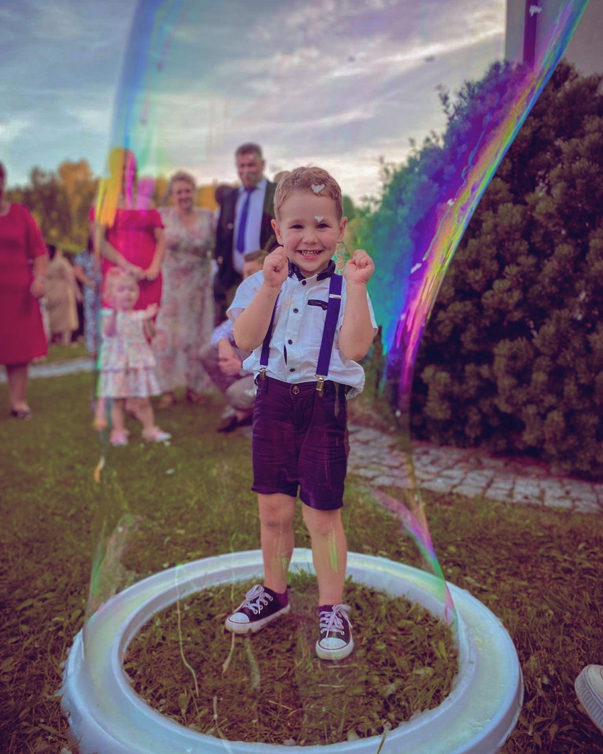 Uśmiechnięty chłopiec w szelkach stoi wewnątrz gigantycznej bańki mydlanej na trawie, w tle goście uroczystości. Kolorowe refleksy na bańce.