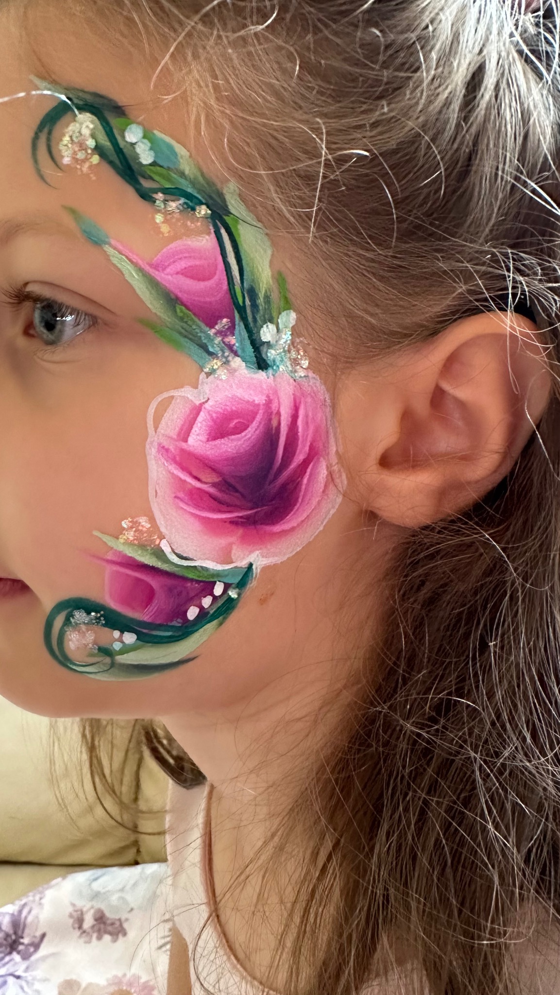 Dziewczynka z artystycznym makijażem twarzy, przedstawiającym różę z zielonymi pędami i białymi akcentami. Widoczny profil twarzy i fragment włosów.