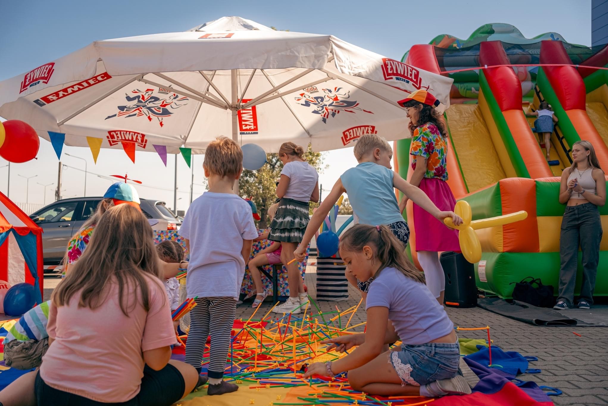 Dzieci bawią się kolorowymi patyczkami na kocu, w tle dmuchany zamek i animator pod parasolem z logo Żywiec. Impreza plenerowa dla dzieci.