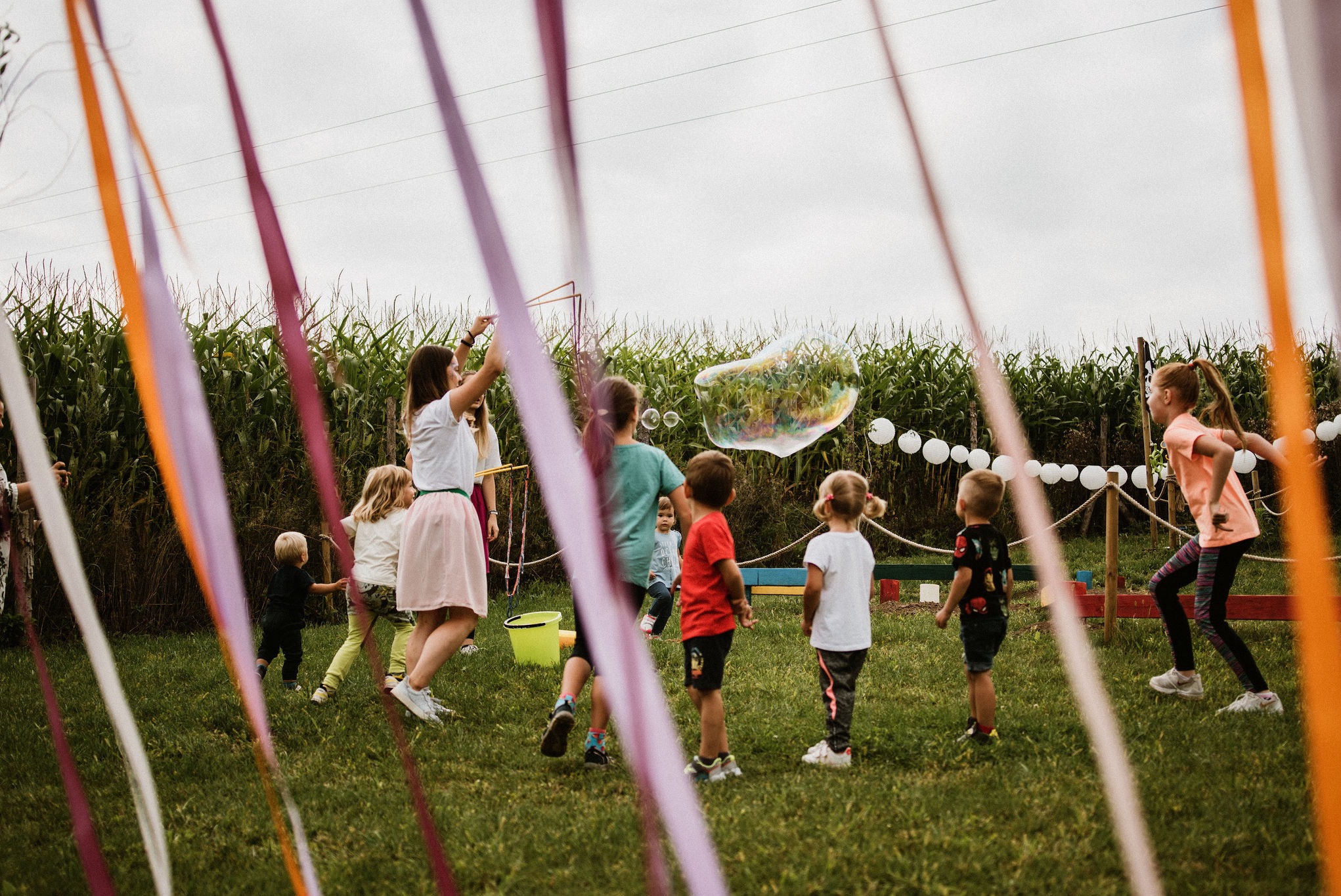 Dzieci bawią się na trawie, łapiąc bańki mydlane podczas animacji na świeżym powietrzu. Kolorowe wstążki i dekoracje tworzą radosną atmosferę.