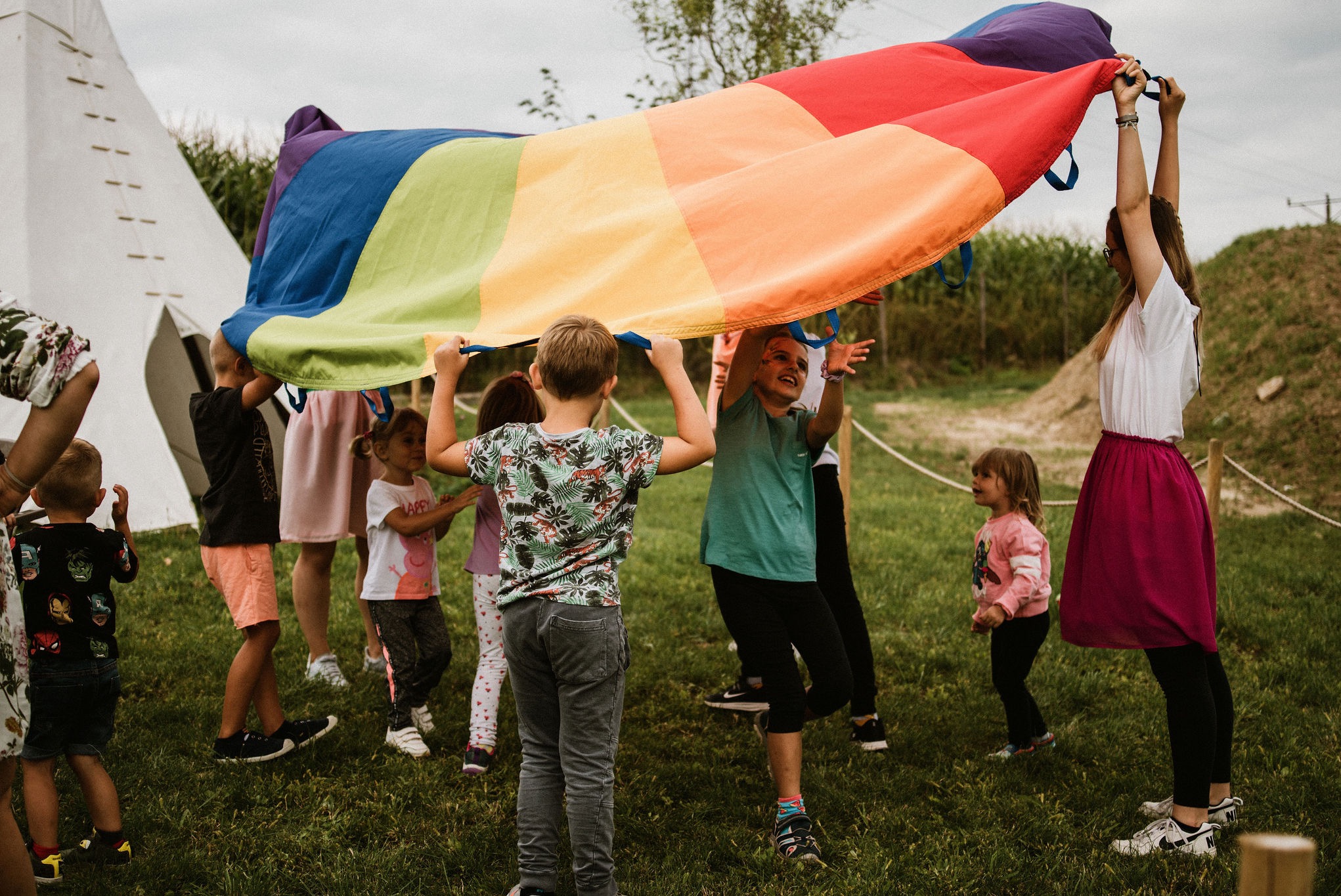 Grupa dzieci bawi się na trawie pod tęczową chustą animacyjną, trzymaną przez animatorkę. W tle białe tipi i zieleń. Radosna atmosfera zabawy na świeżym powietrzu.