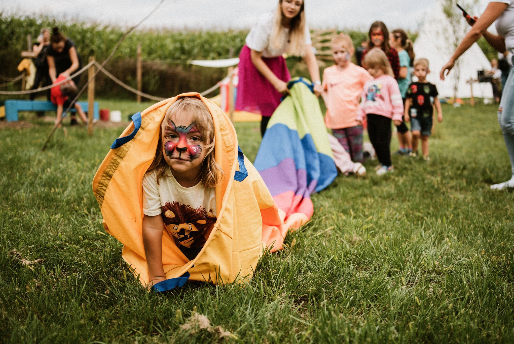 Dziecko z pomalowaną twarzą wychodzi z tunelu z materiału na trawie. W tle grupa dzieci i animatorzy podczas zabawy z kolorową chustą animacyjną.