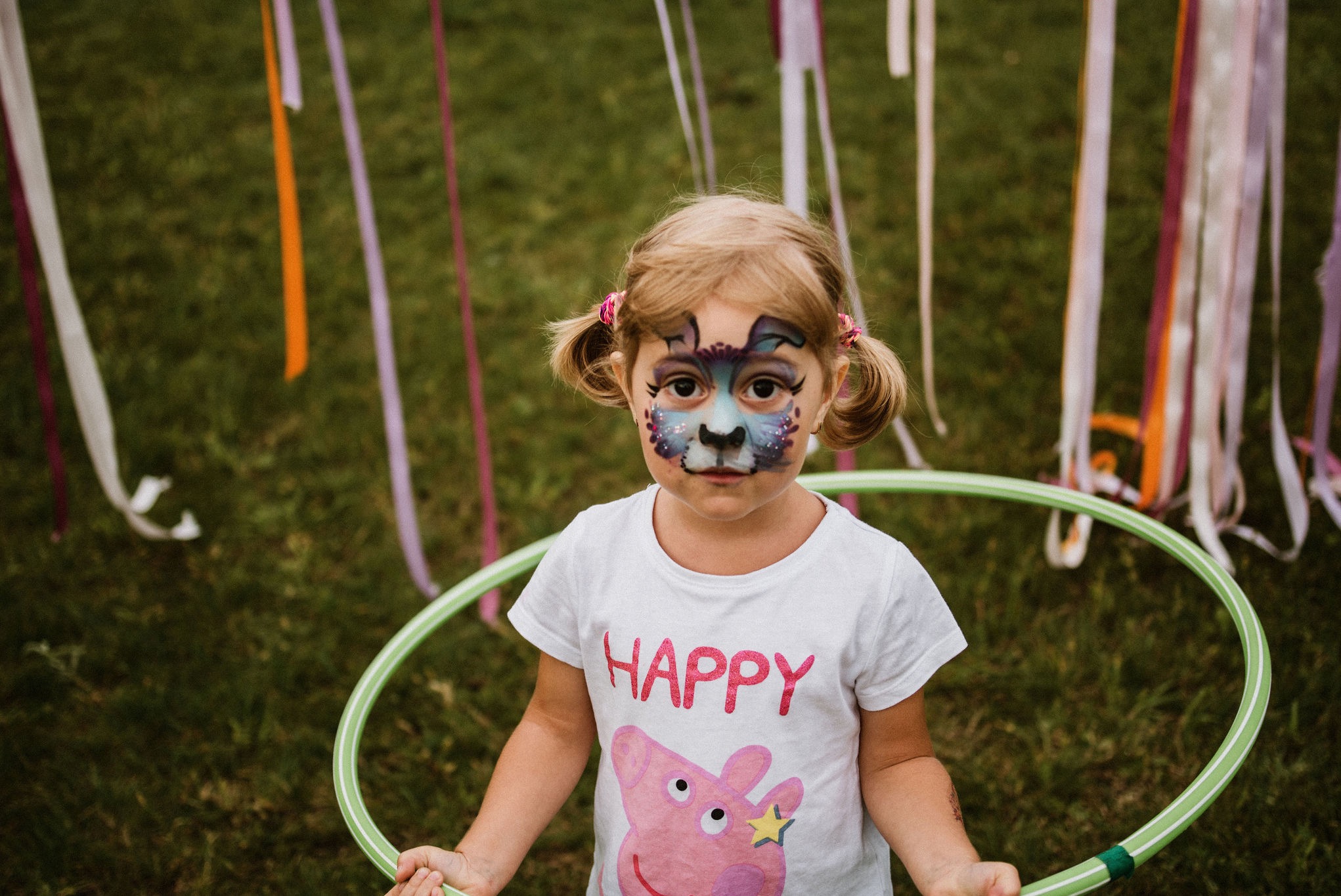Dziewczynka z pomalowaną twarzą w motywy zwierzęce, trzymająca zielone koło hula-hop na tle trawy i kolorowych wstążek. Uśmiech na twarzy, radosna atmosfera.