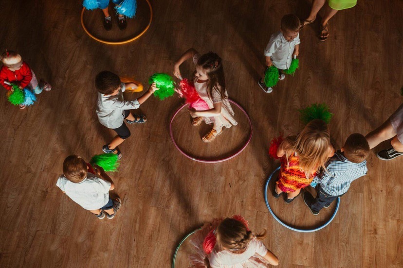 Zabawa z hula-hop i pomponami z perspektywy lotu ptaka. Dzieci w różnym wieku bawią się na drewnianej podłodze. Kolorowe rekwizyty i radosna atmosfera.
