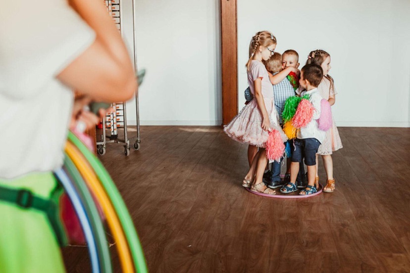 Grupa dzieci otoczona kolorowymi pomponami i hula-hop, stojąca na fioletowym kole. W tle jasne ściany i drewniana podłoga. Ujęcie z boku, z widocznym fragmentem osoby dorosłej.
