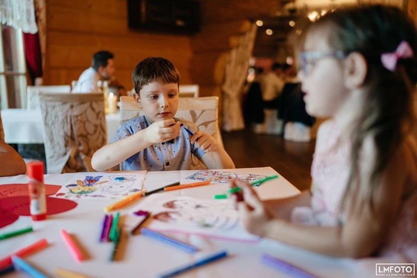 Dzieci rysują kolorowe obrazki przy stole w drewnianym wnętrzu. Widoczne kredki, kartki i klej. Uczestnicy imprezy w tle.