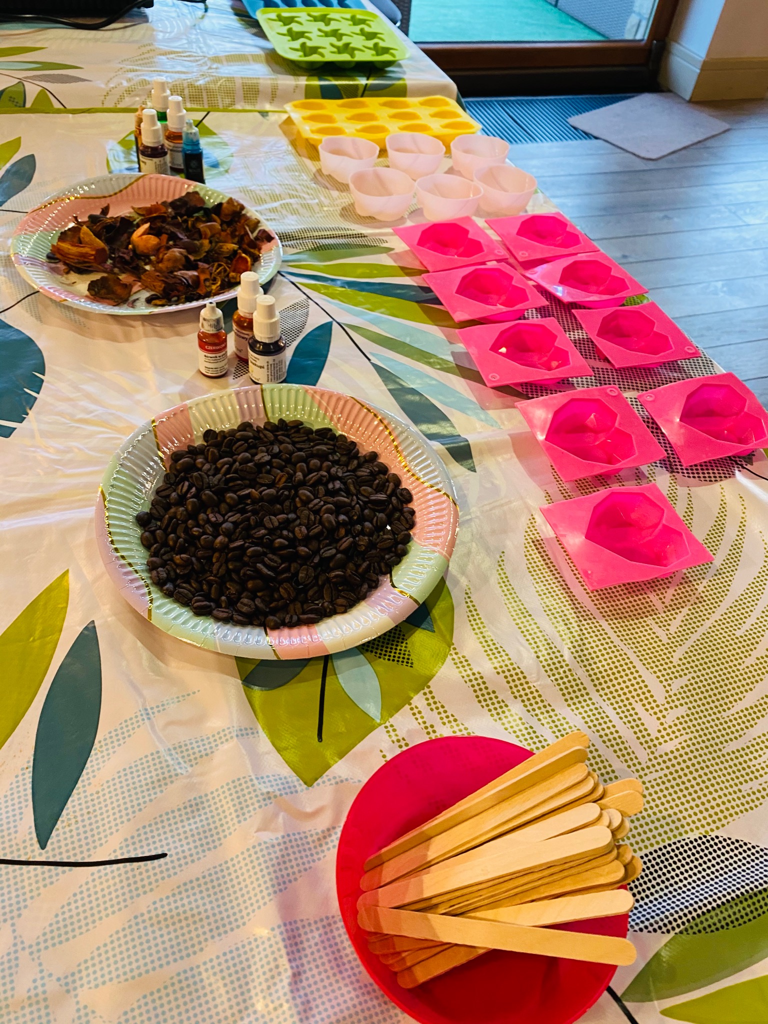 Przygotowane składniki: ziarna kawy, suszone kwiaty, barwniki, foremki i patyczki na stole z wzorzystym obrusem, sugerujące warsztaty rękodzielnicze.