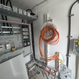 Filip Dytman - Otwarta skrzynka elektryczna z widocznymi bezpiecznikami i okablowaniem. Pomarańczowe i białe przewody zwisają ze ściany. Widoczne oznaczenia garaży na półce.