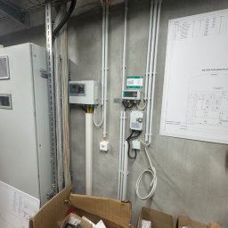 Filip Dytman - Szara ściana z zamontowanymi przewodami elektrycznymi w białych rurkach, skrzynka elektryczna i schemat instalacji. Na podłodze kartony z elementami instalacji.