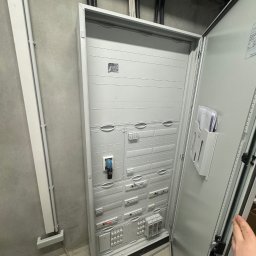 Filip Dytman - Otwarta, szara szafa elektryczna z widocznymi zabezpieczeniami i elementami instalacji w surowym wnętrzu. Widoczne kable i rury instalacyjne na ścianie i suficie.