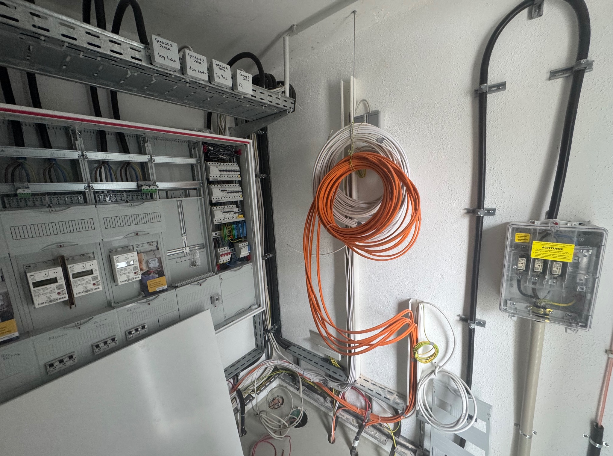 Otwarta skrzynka elektryczna z widocznymi bezpiecznikami i okablowaniem. Pomarańczowe i białe przewody zwisają ze ściany. Widoczne oznaczenia garaży na półce.