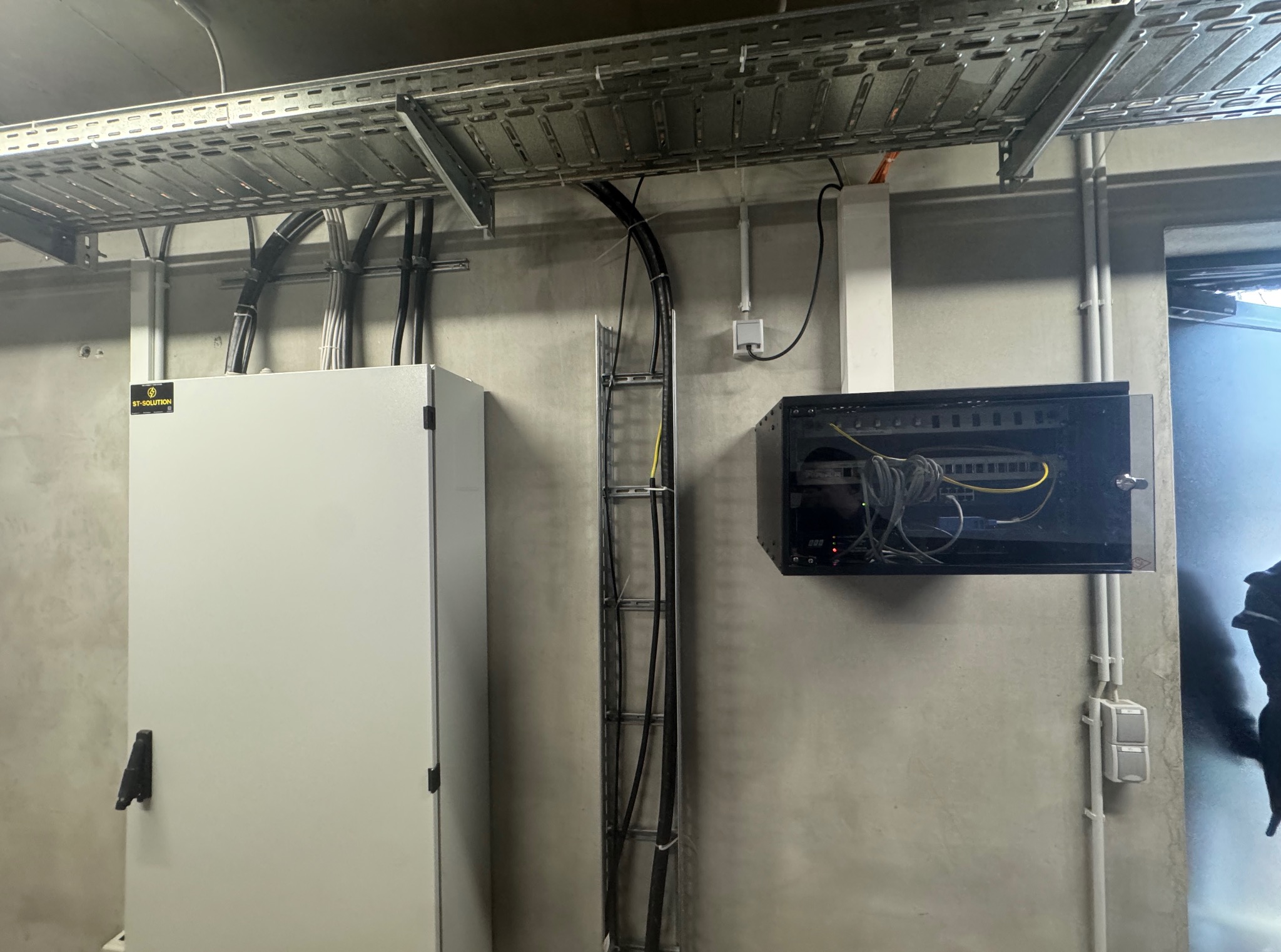 Szara ściana z metalowymi korytkami kablowymi pod sufitem, szafa sterownicza z logo, pionowa drabinka kablowa i szafka RACK z przewodami. Widok na instalacje wewnątrz budynku.