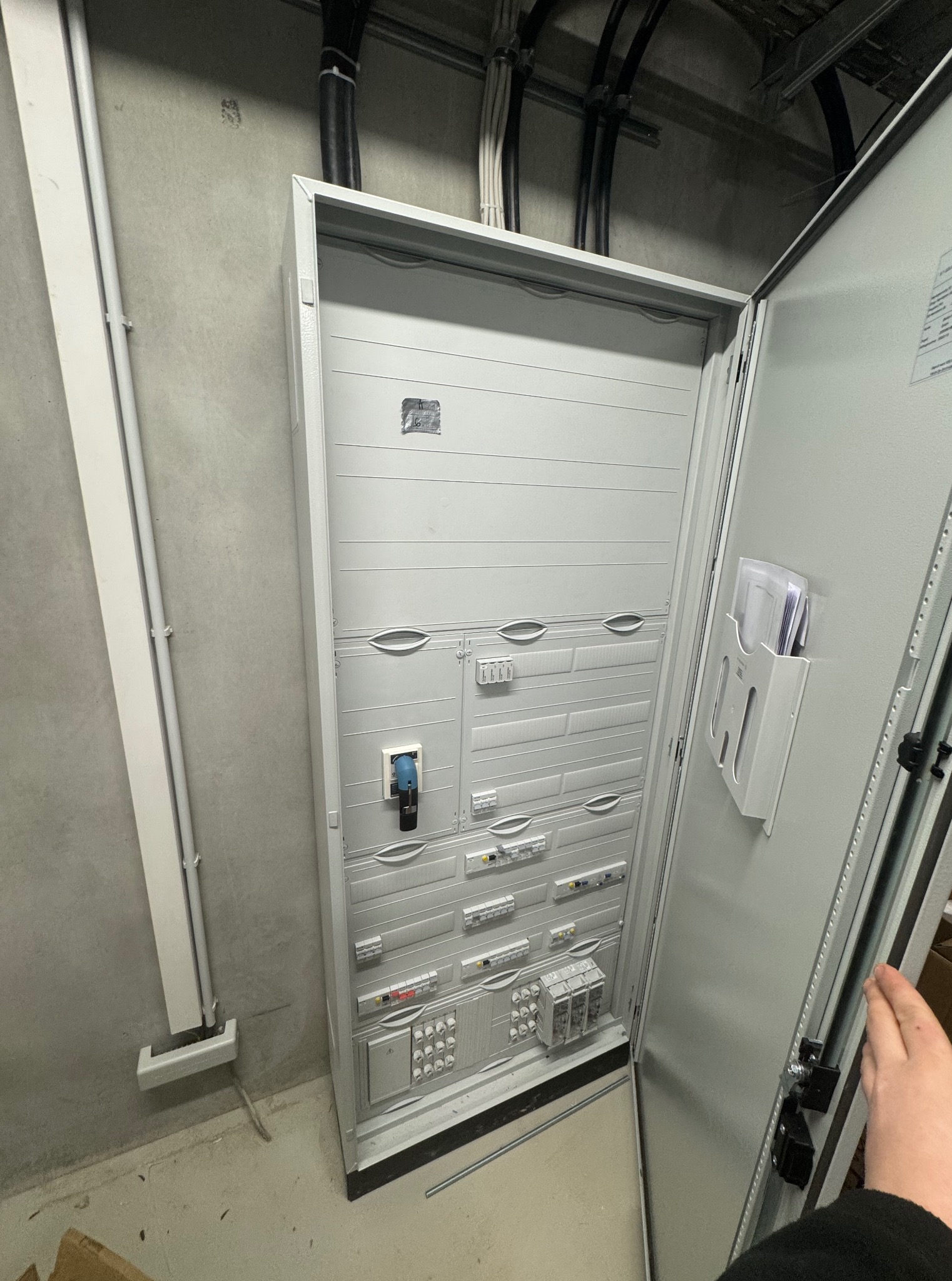 Otwarta, szara szafa elektryczna z widocznymi zabezpieczeniami i elementami instalacji w surowym wnętrzu. Widoczne kable i rury instalacyjne na ścianie i suficie.