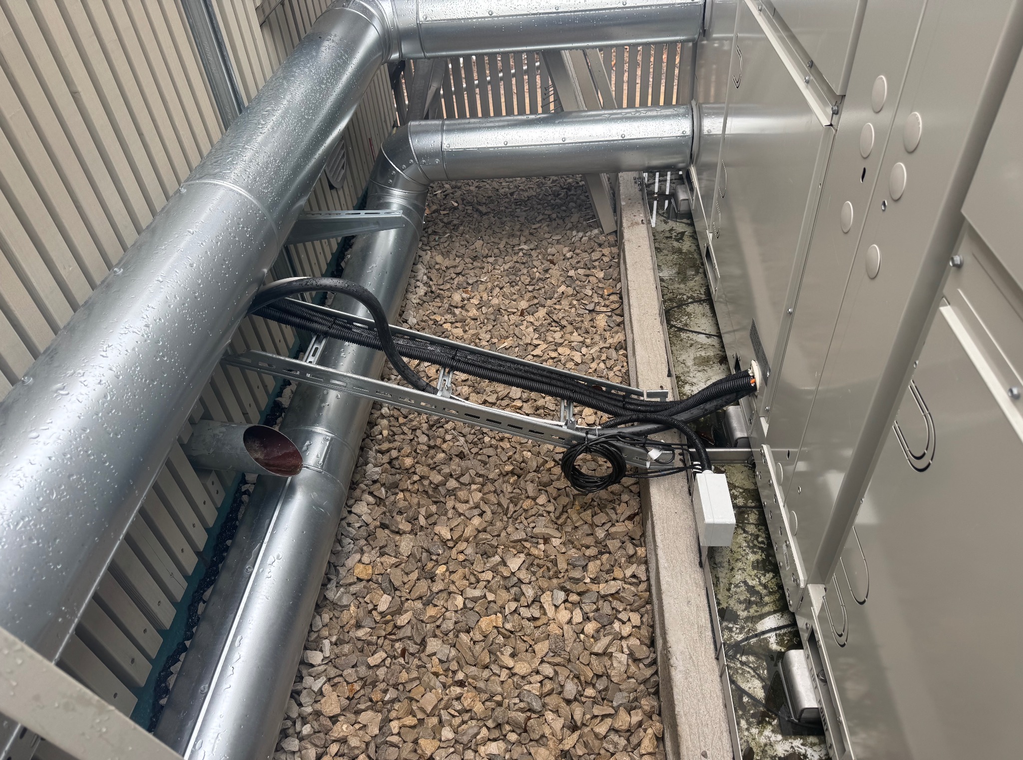 Metalowe rury wentylacyjne i przewody elektryczne na żwirowym podłożu, zamontowane na zewnątrz budynku. Widoczne elementy instalacji i skrzynka elektryczna.