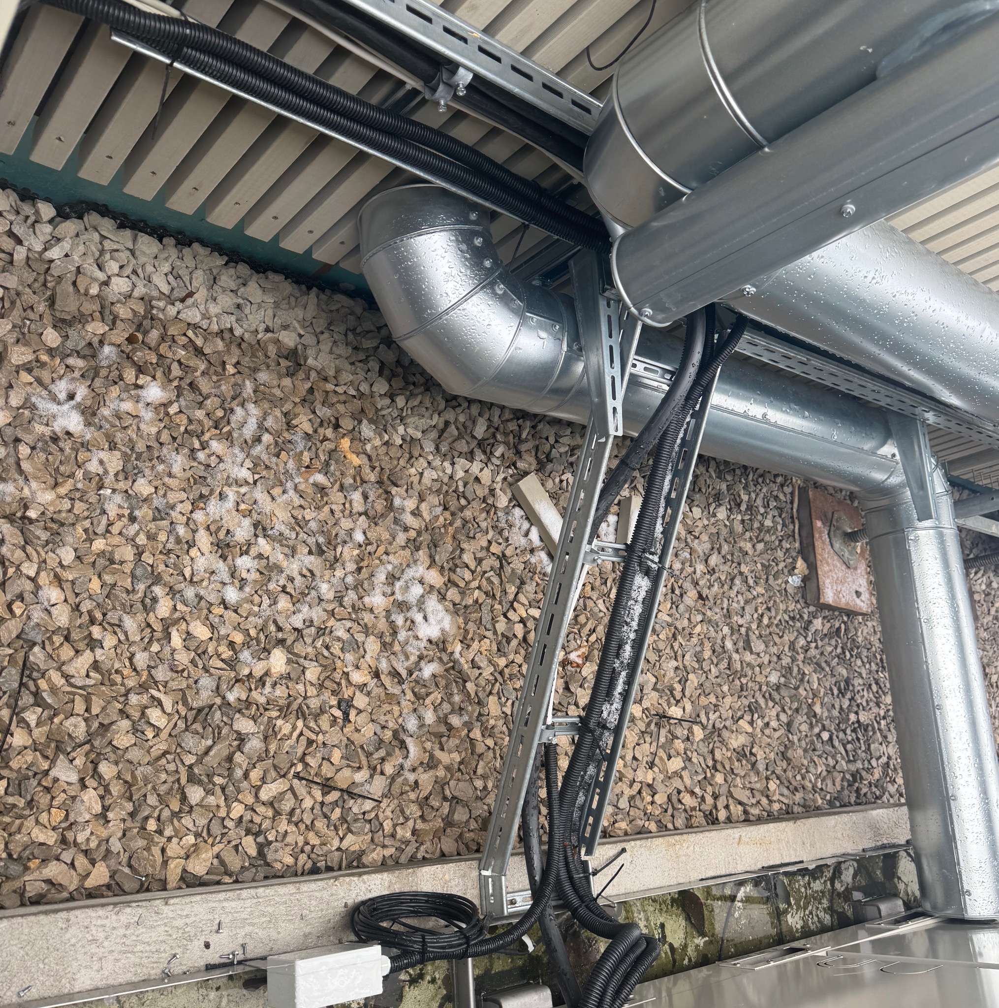 Metalowe rury wentylacyjne i czarne kable w peszlach, zamontowane na zewnątrz budynku, widoczne na tle ściany z kamieni. Elementy instalacji na wspornikach.