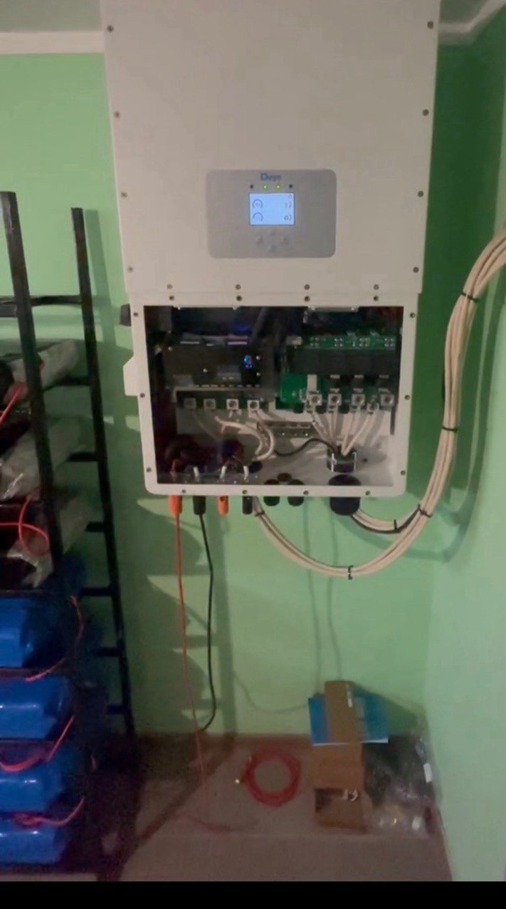 Zamontowany inwerter solarny z otwartą obudową, widoczne okablowanie i podłączenia. Baterie akumulatorowe obok drabiny. Ściana w odcieniach zieleni.