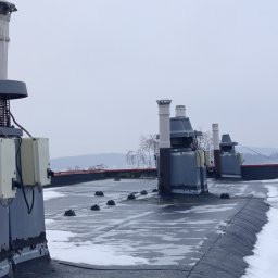 ELEKTRO AL SPÓŁKA Z OGRANICZONĄ ODPOWIEDZIALNOŚCIĄ - Szary dach pokryty śniegiem z widocznymi elementami instalacji odgromowej i wentylacyjnej. Na pierwszym planie skrzynki elektryczne. Pochmurne niebo w tle.