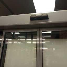 ELEKTRO AL SPÓŁKA Z OGRANICZONĄ ODPOWIEDZIALNOŚCIĄ - Automatyczne drzwi przesuwne z czujnikiem GEZE, widoczne wewnątrz budynku. Szklane panele odbijają oświetlenie sufitowe i wnętrze sklepu.