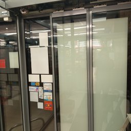 ELEKTRO AL SPÓŁKA Z OGRANICZONĄ ODPOWIEDZIALNOŚCIĄ - Automatyczne drzwi przesuwne z widocznym mechanizmem napędowym i naklejkami informacyjnymi na szkle. Widoczny fragment drabiny.