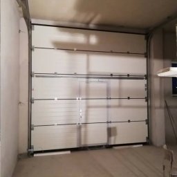 ELEKTRO AL SPÓŁKA Z OGRANICZONĄ ODPOWIEDZIALNOŚCIĄ - Biała segmentowa brama garażowa z drzwiami wejściowymi, zamontowana w surowym wnętrzu garażu. Widoczne prowadnice i elementy mocujące.