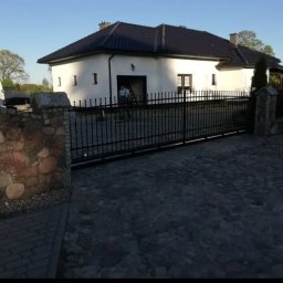 ELEKTRO AL SPÓŁKA Z OGRANICZONĄ ODPOWIEDZIALNOŚCIĄ - Czarna, metalowa brama wjazdowa z automatyką, zamontowana przed białym domem z ciemnym dachem. Kamienne słupki i podjazd z kostki brukowej. Widoczny monter.