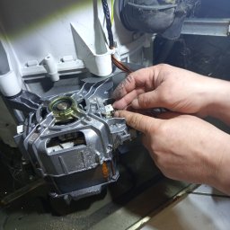 ELEKTRO AL SPÓŁKA Z OGRANICZONĄ ODPOWIEDZIALNOŚCIĄ - Silnik pralki w trakcie naprawy: dłonie serwisanta podłączają przewody do elementu. Widoczne zabrudzenia i pajęczyny wewnątrz urządzenia.