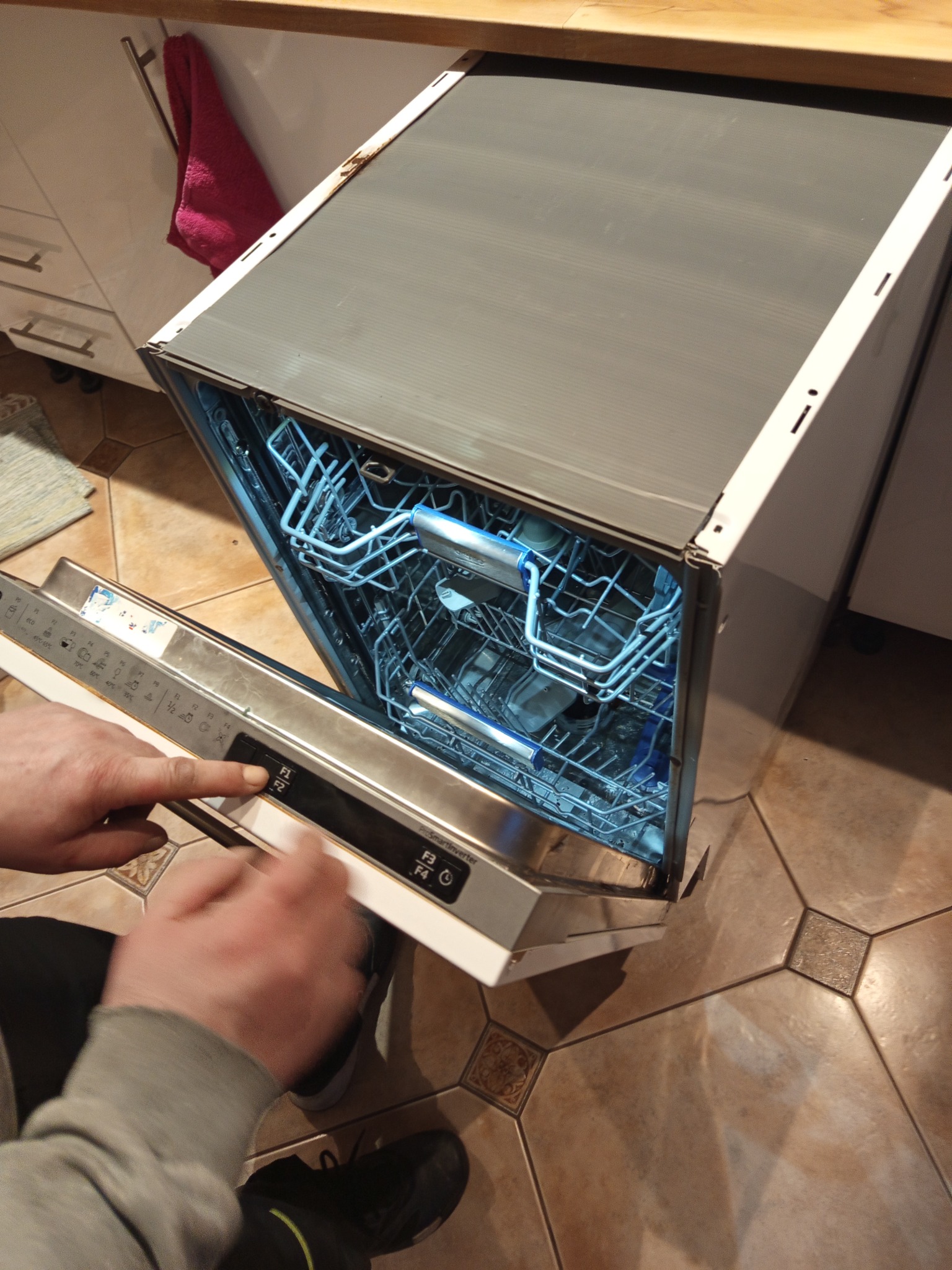 Otwarta zmywarka z podświetlonym wnętrzem, dłoń naciska przycisk na panelu sterowania. Widoczna zabudowa kuchenna i fragment podłogi z płytek.