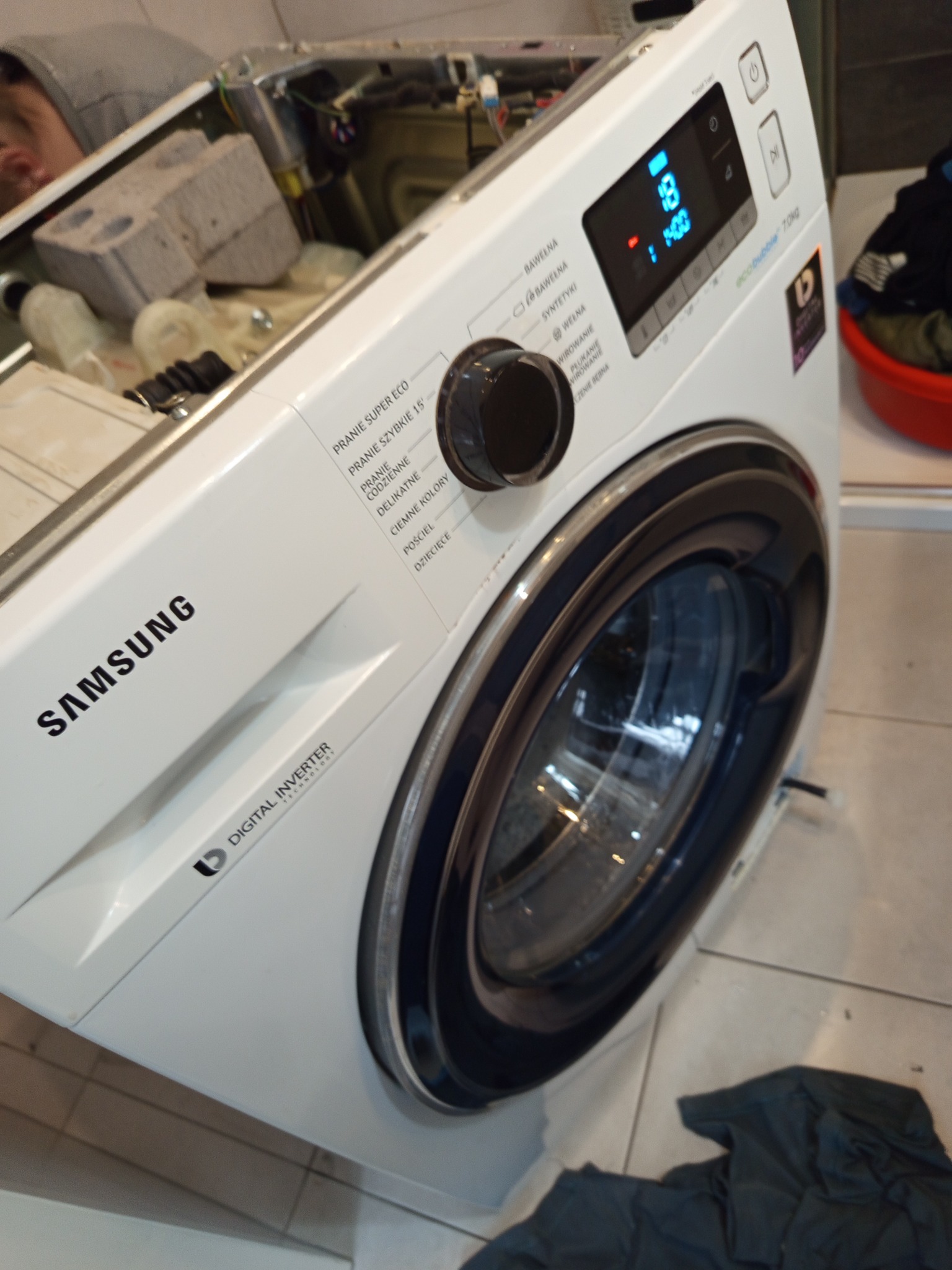 Otwarta pralka Samsung podczas naprawy, widoczne wnętrze i panel sterowania. Ujęcie z bliska, lekko z góry, z widocznymi elementami wnętrza i częścią osoby.