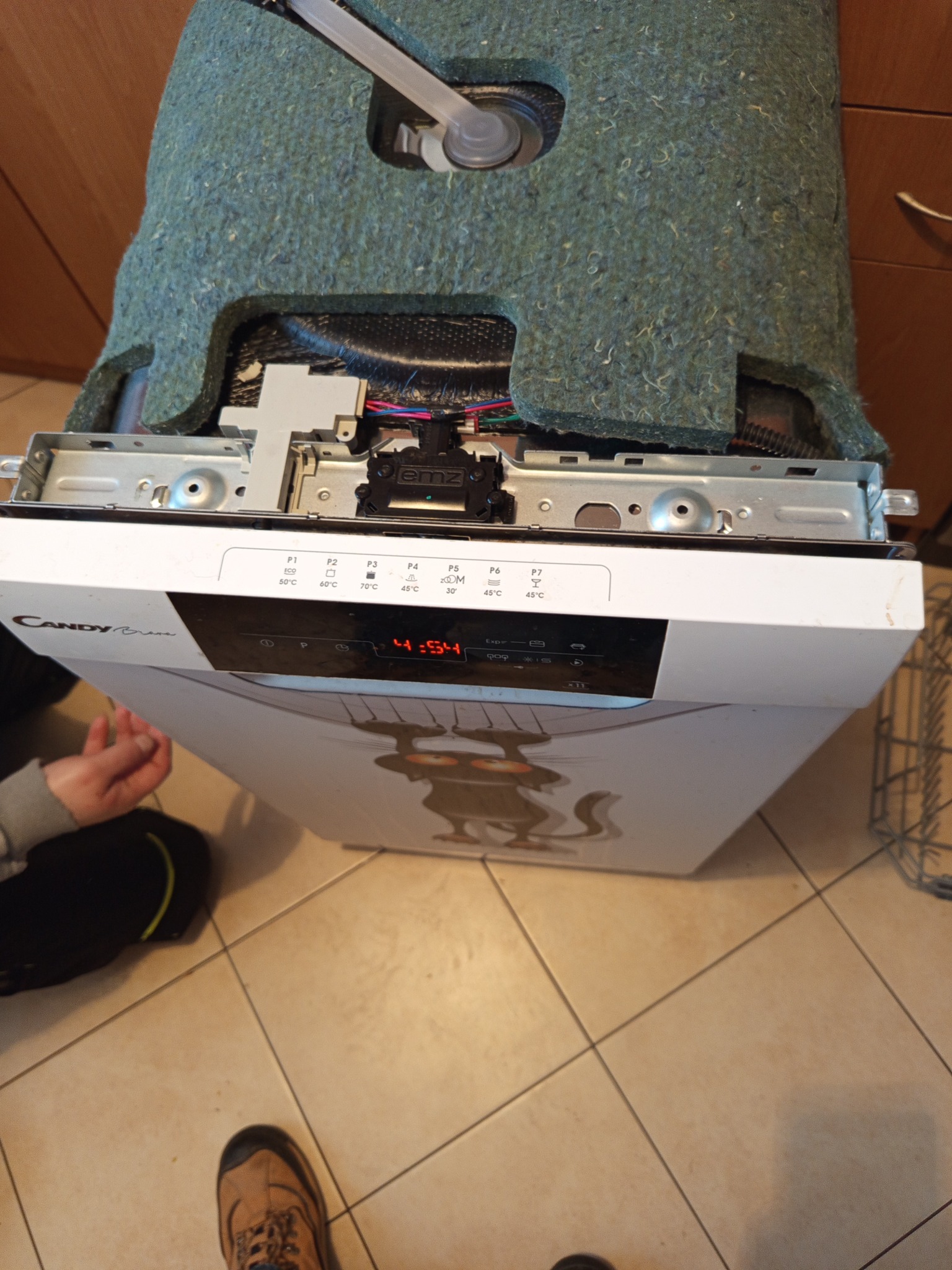 Otwarta zmywarka Candy z widocznymi elementami wewnętrznymi i panelem sterowania, ustawiona na kafelkach. Widoczne ręce osoby i fragmenty obuwia.