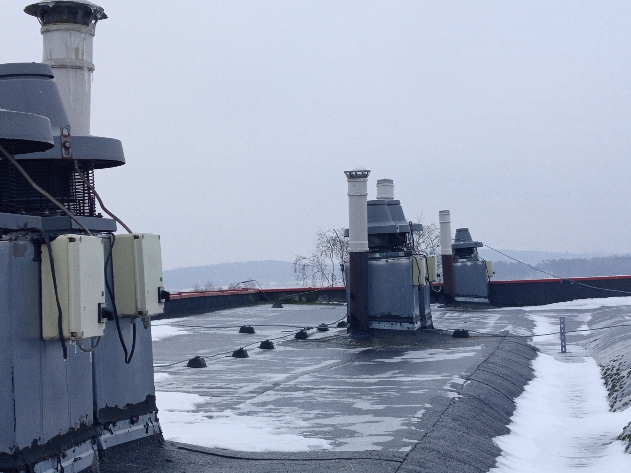 Szary dach pokryty śniegiem z widocznymi elementami instalacji odgromowej i wentylacyjnej. Na pierwszym planie skrzynki elektryczne. Pochmurne niebo w tle.