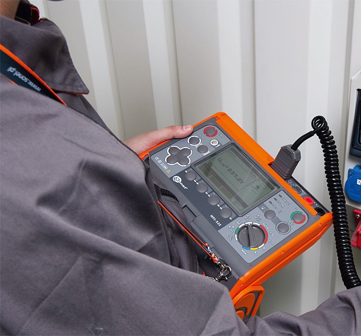 Elektryk w szarym kombinezonie mierzy napięcie miernikiem Sonel MPI-525 przy gniazdku elektrycznym. Widoczny przewód spiralny i fragment ściany z panelami.