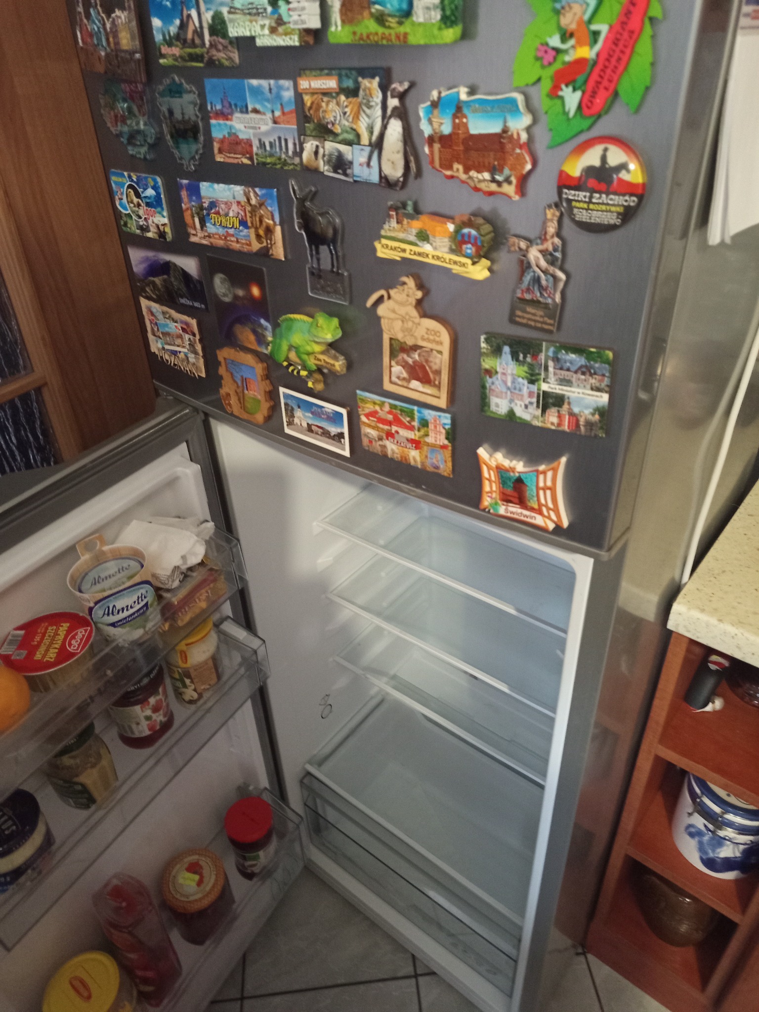 Otwarta lodówka z magnesami pamiątkowymi na drzwiach, półki częściowo wypełnione produktami spożywczymi. Widoczne słoiki, jogurty i inne artykuły spożywcze.