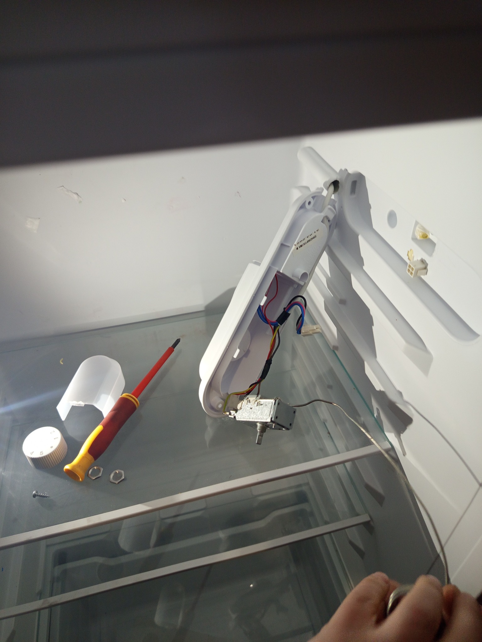 Wnętrze lodówki w trakcie naprawy w Liśnicy. Widoczne elementy elektryczne, śrubokręt i regulator temperatury na szklanej półce. Ujęcie z góry, oświetlenie dzienne.