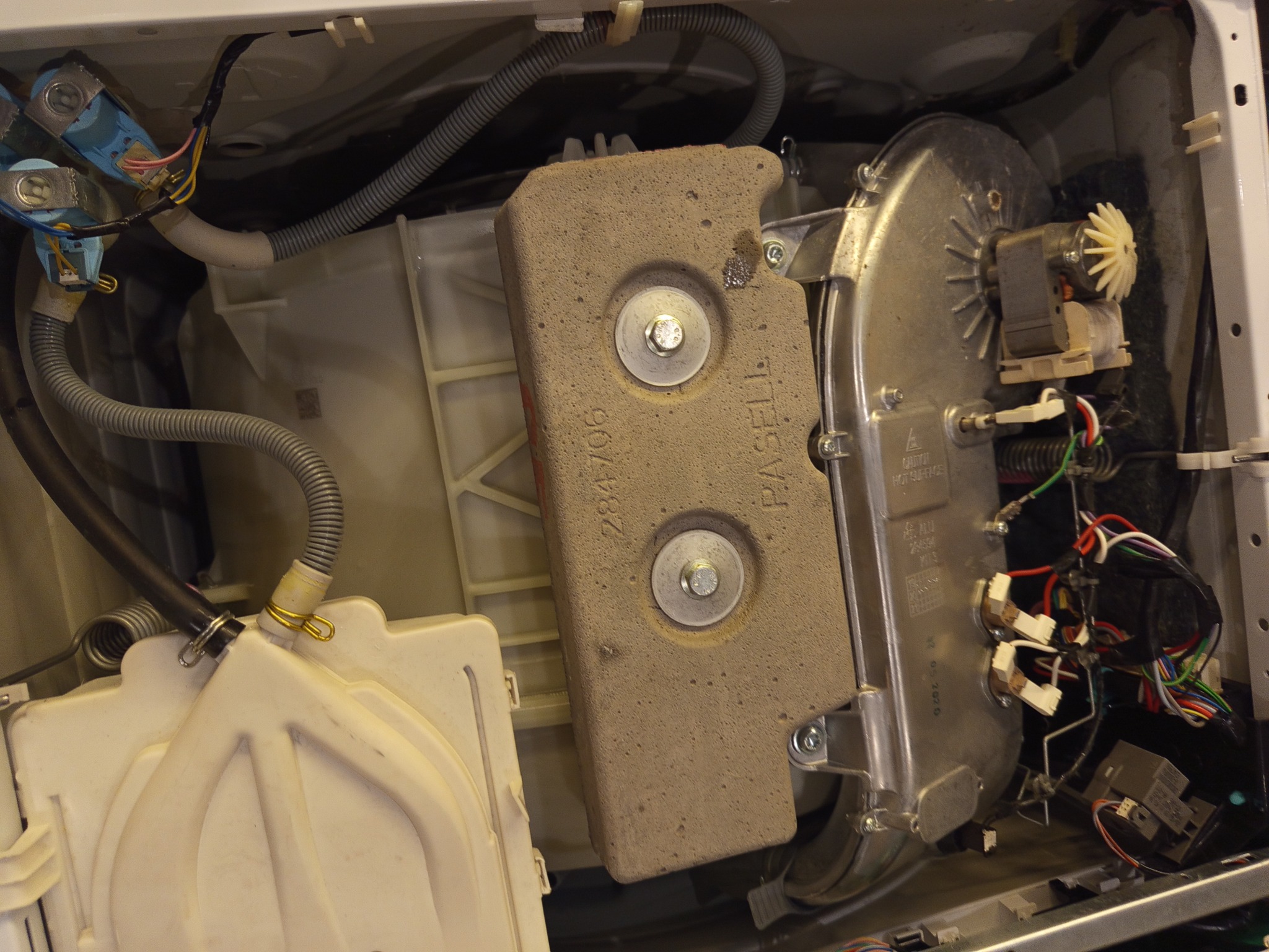 Wnętrze pralki z widocznymi elementami grzałki, przewodami i amortyzatorem. Detaliczne ujęcie na podzespoły AGD w trakcie naprawy lub serwisu.