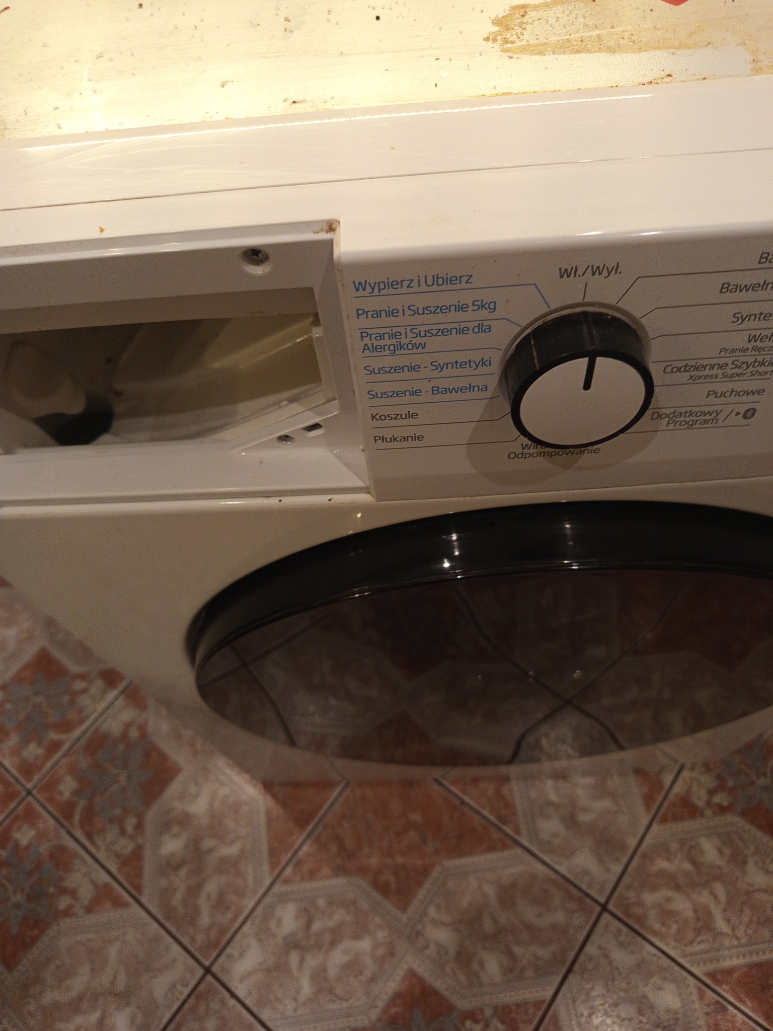 Biała pralka z otwartą szufladą na detergent i widocznym panelem sterowania z programami prania, ustawiona na wzorzystej, brązowej podłodze.