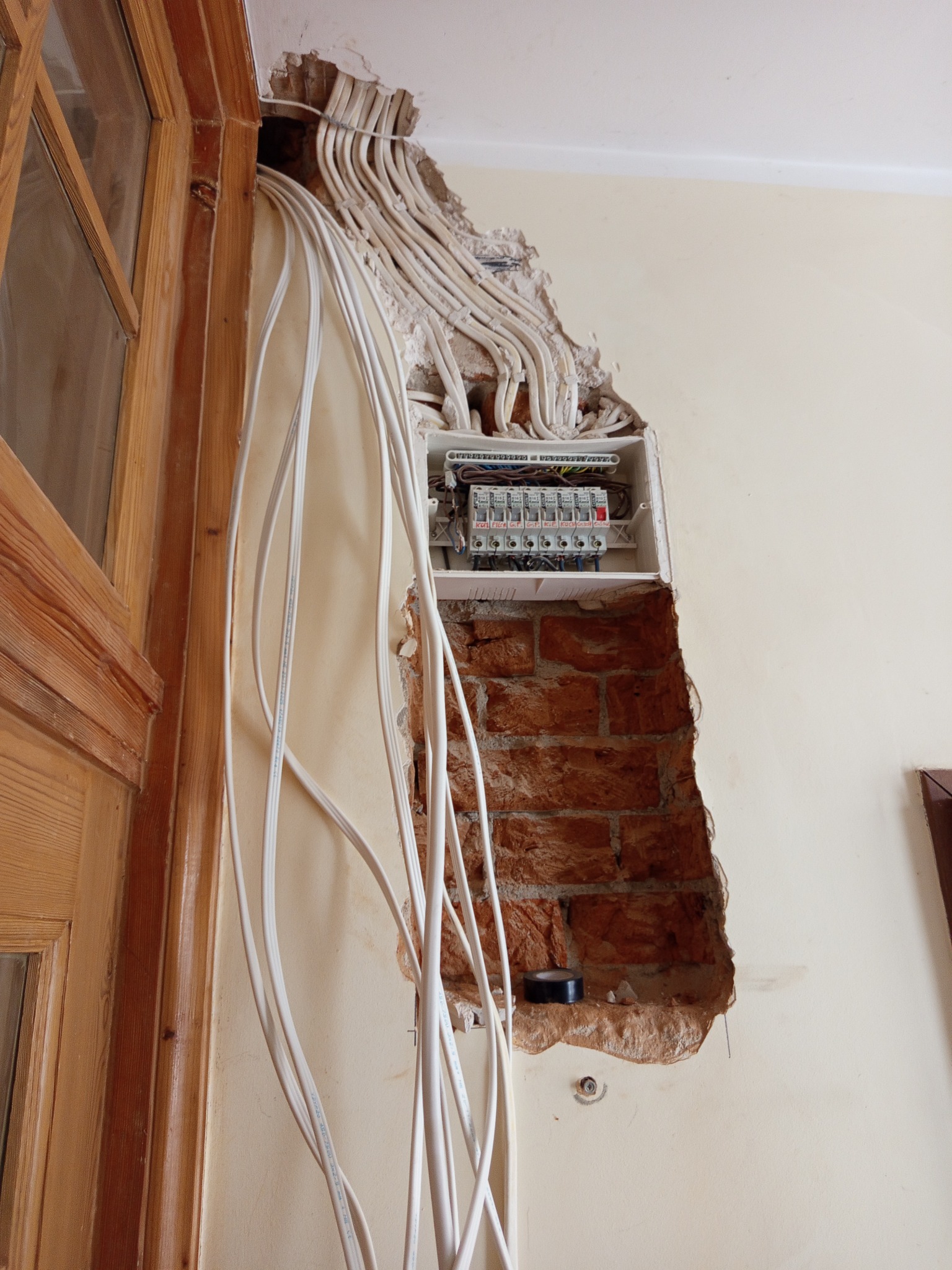 Otwarta puszka elektryczna w ścianie z widocznymi przewodami, obok framuga drzwi. Widoczne ślady po kuciu w ścianie z cegły.