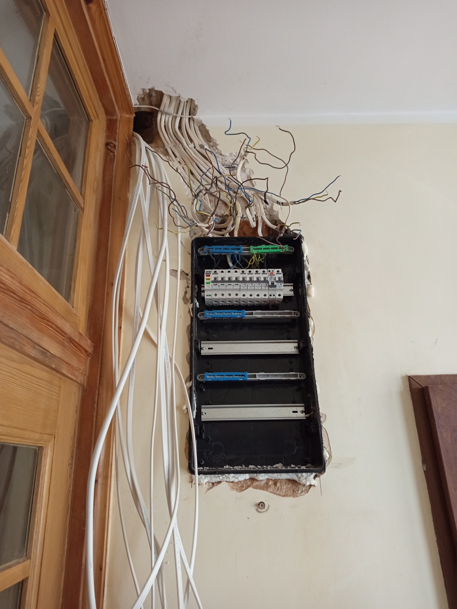 Otwarta skrzynka elektryczna w ścianie z wystającymi kablami, widoczne bezpieczniki i listwy montażowe. Prace instalacyjne w trakcie, obok drewniana futryna drzwi.