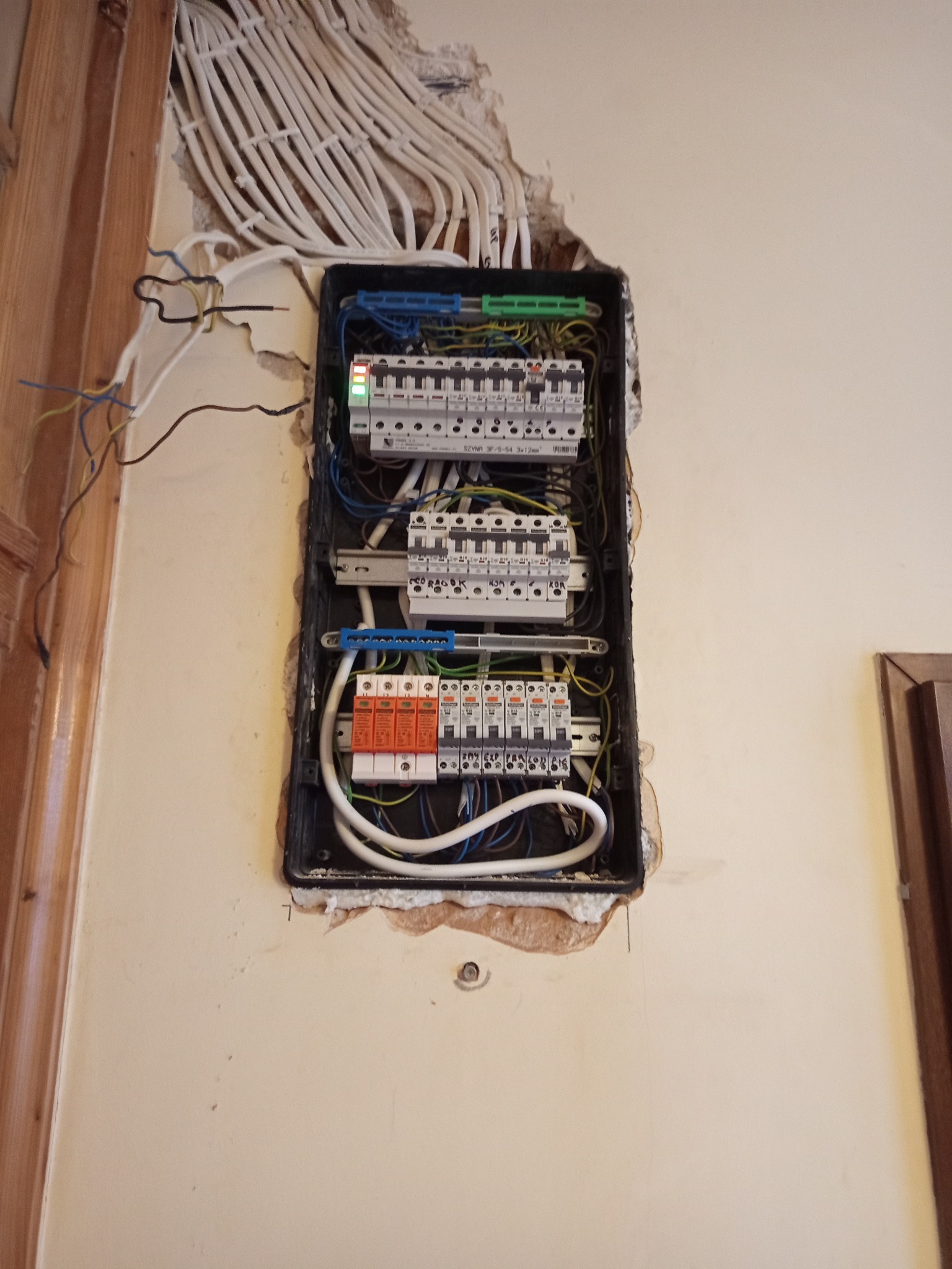 Otwarta skrzynka bezpiecznikowa z widocznymi przewodami i zabezpieczeniami, zamontowana na ścianie z uszkodzonym tynkiem. Widoczne kable wchodzące do skrzynki.