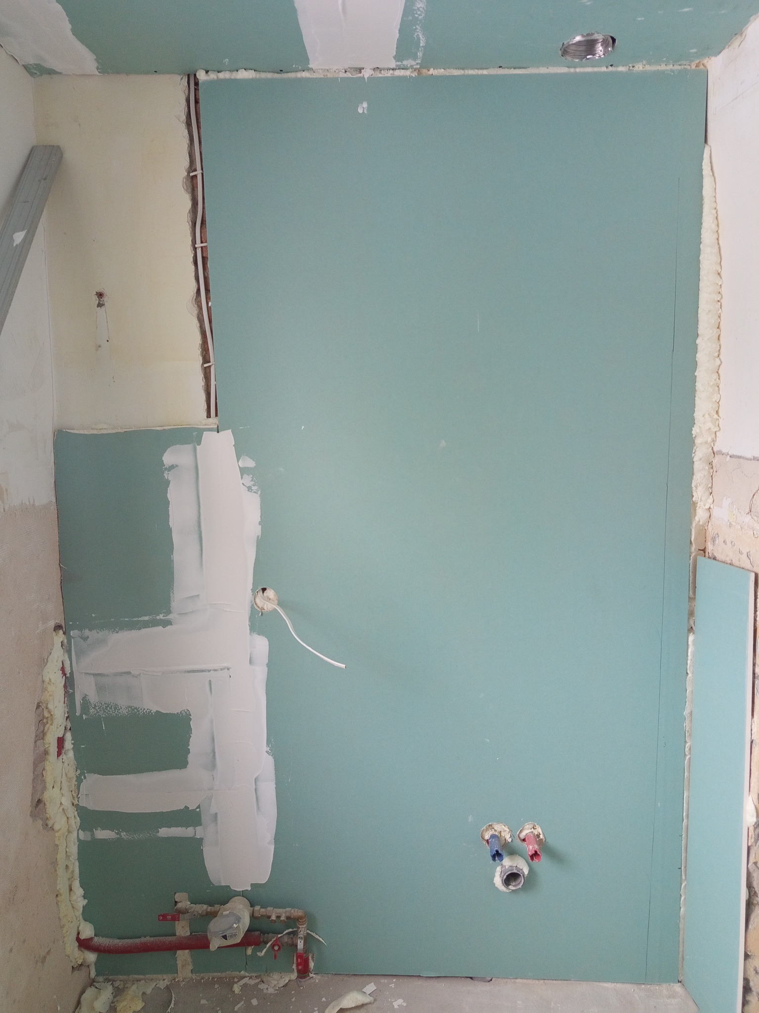 Montaż płyt gipsowo-kartonowych na ścianie z widocznymi instalacjami elektrycznymi i hydraulicznymi. Częściowo zagruntowana powierzchnia, pianka montażowa w szczelinach.