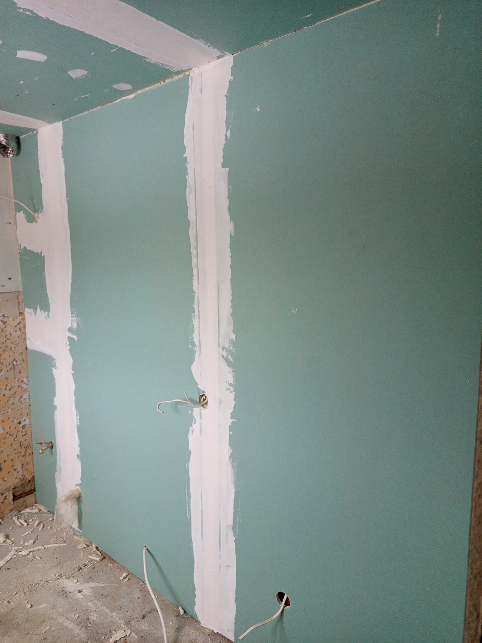 Ściana z płyt gipsowo-kartonowych w trakcie szpachlowania, widoczne kable elektryczne wystające ze ściany. Prace wykończeniowe w surowym wnętrzu.