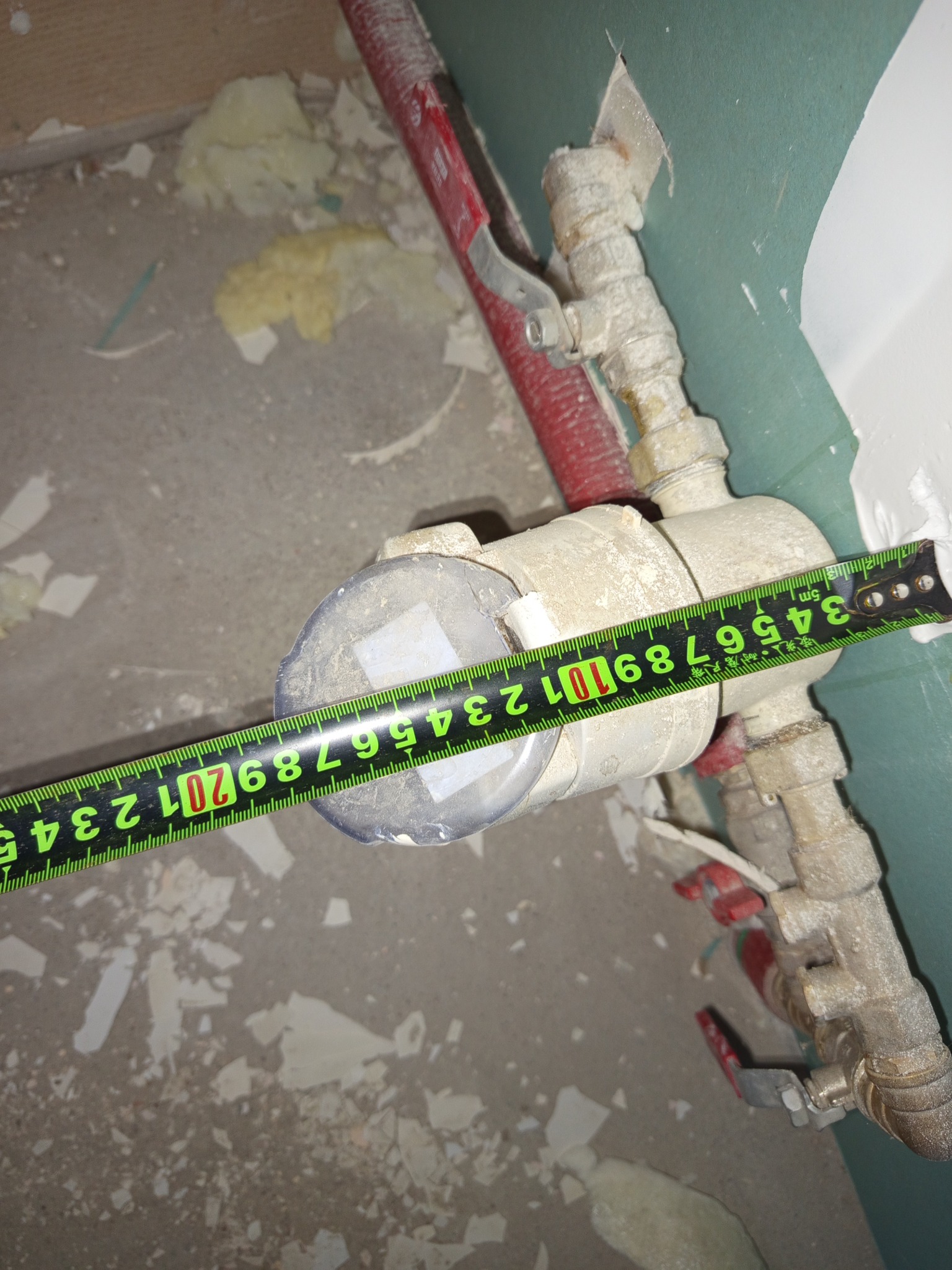 Brudny wodomierz z taśmą mierniczą na tle ściany i podłogi z resztkami materiałów budowlanych. Widoczne rury i zawory.