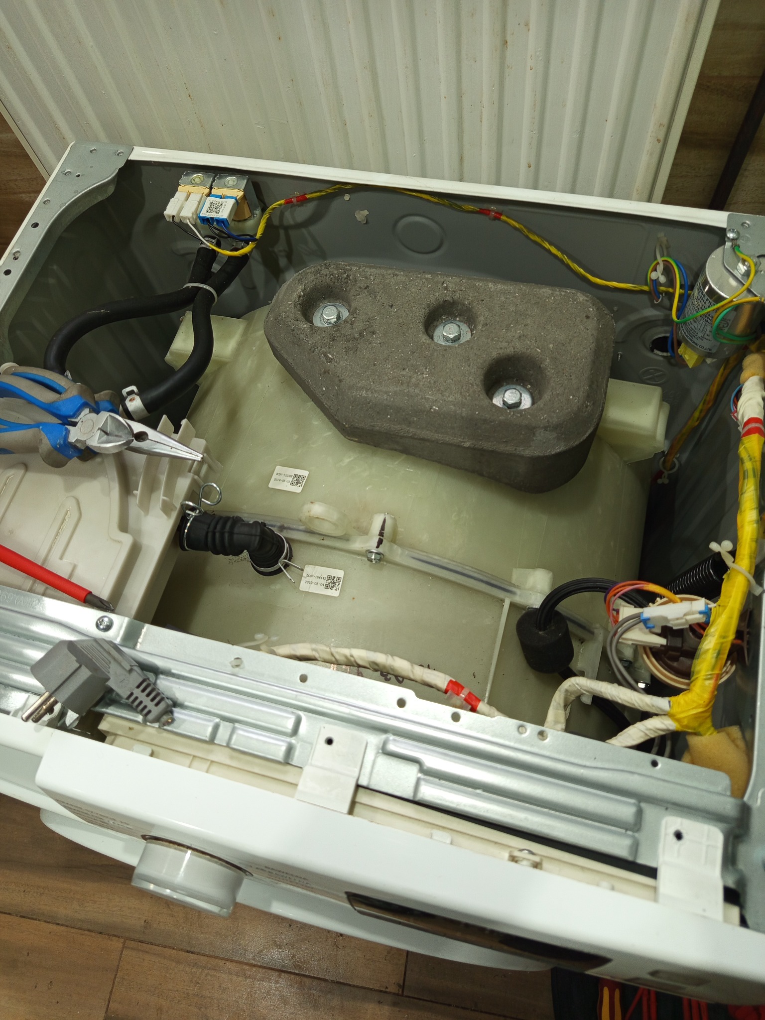 Wnętrze rozmontowanej pralki z widocznymi elementami: bęben, obciążnik betonowy, przewody elektryczne i narzędzia. Ukazany etap naprawy lub serwisu AGD.