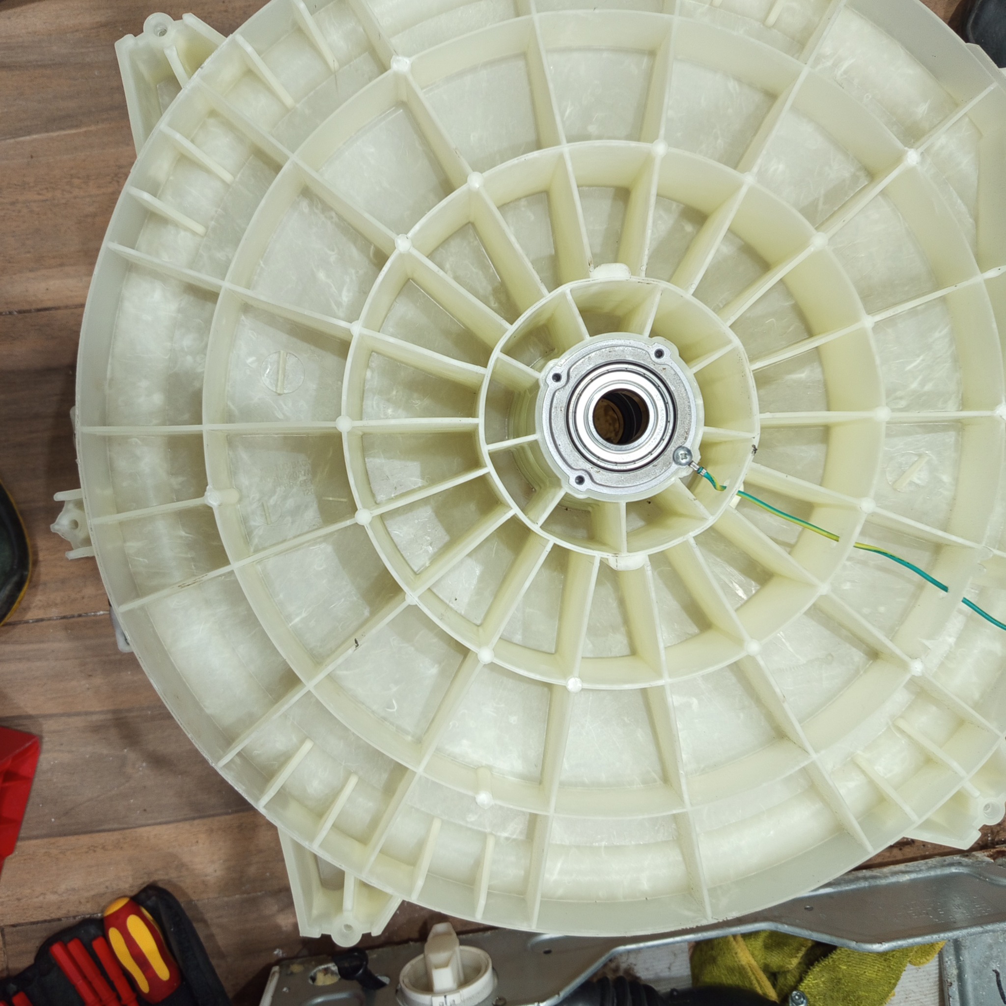 Rozłożony na części bęben pralki automatycznej z widocznym łożyskiem i elementami konstrukcyjnymi, otoczony narzędziami na drewnianej podłodze.