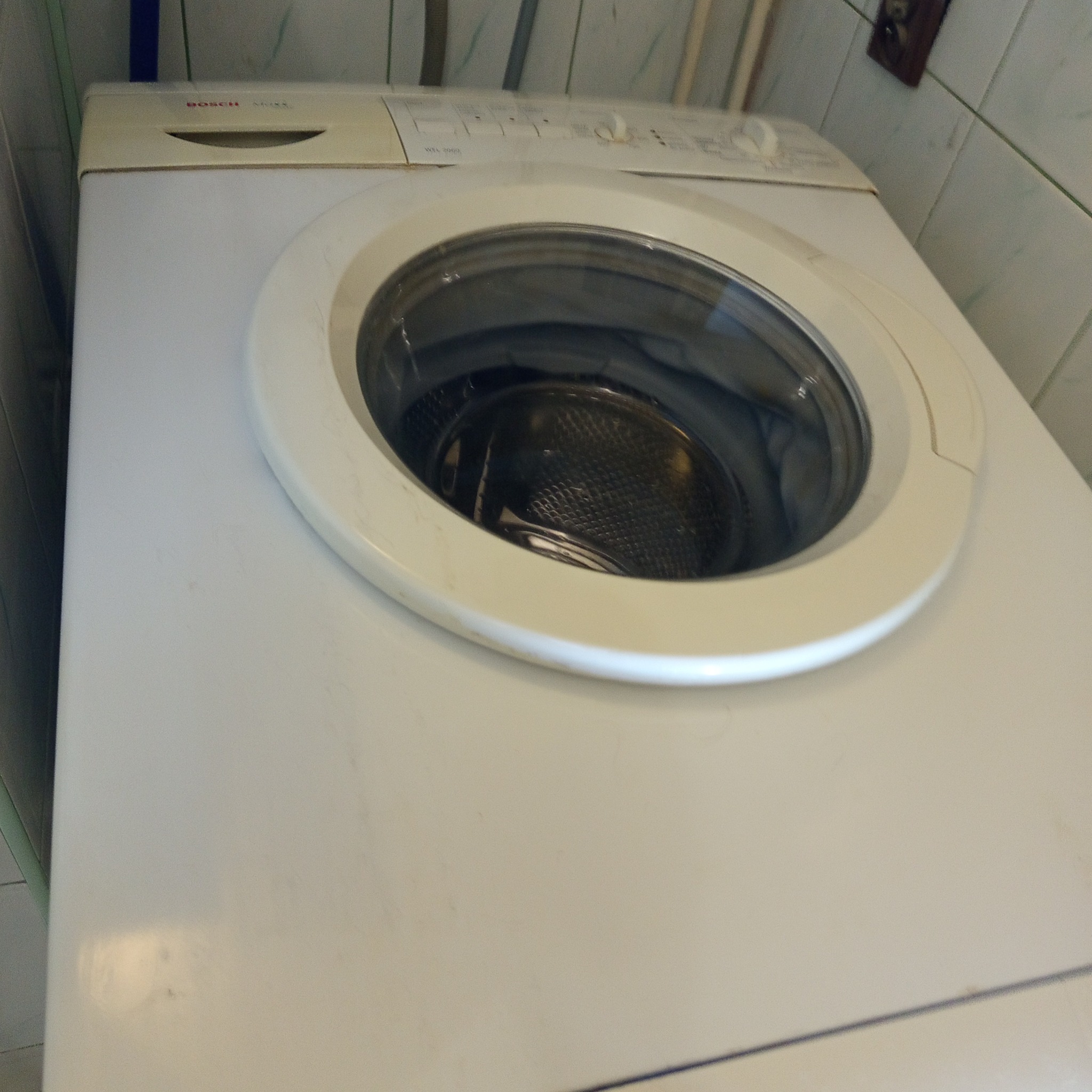 Biała pralka Bosch z widocznym bębnem, ustawiona w łazience z kafelkami. Ujęcie z góry, lekko z boku, ukazujące detale urządzenia i otoczenia.